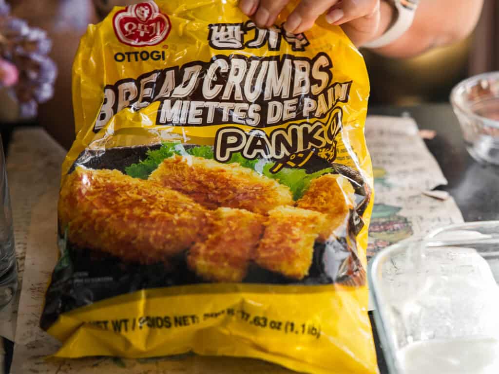 A person holds a yellow bag of Ottogi Panko Bread Crumbs over a table. The packaging shows an image of fried, breaded food. The bag is labeled in English and a foreign language.